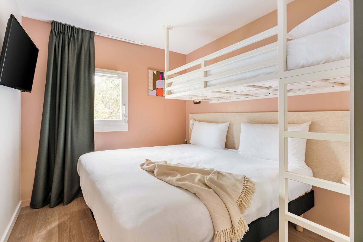 A bedroom with a bunk bed, a flat-screen TV mounted on the wall, a window with curtains, and a bedside table.