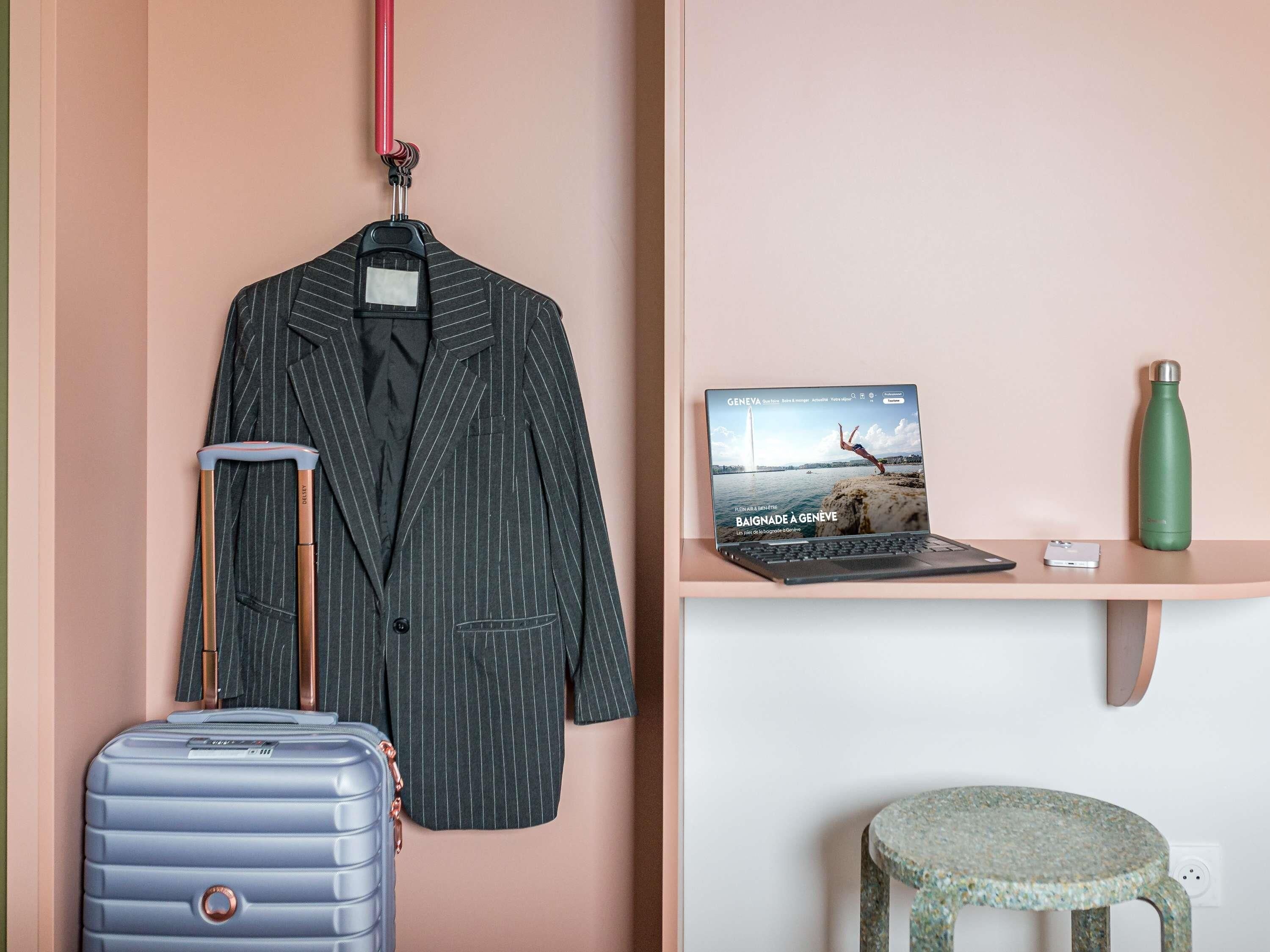 A room with a hanging coat rack, a laptop on a desk, a green water bottle, and a suitcase.
