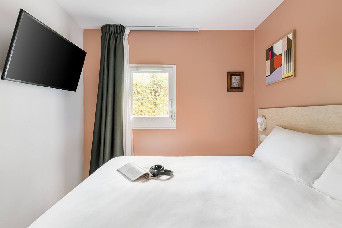A hotel room with a bed, a TV mounted on the wall, a window with curtains, and a painting on the wall.
