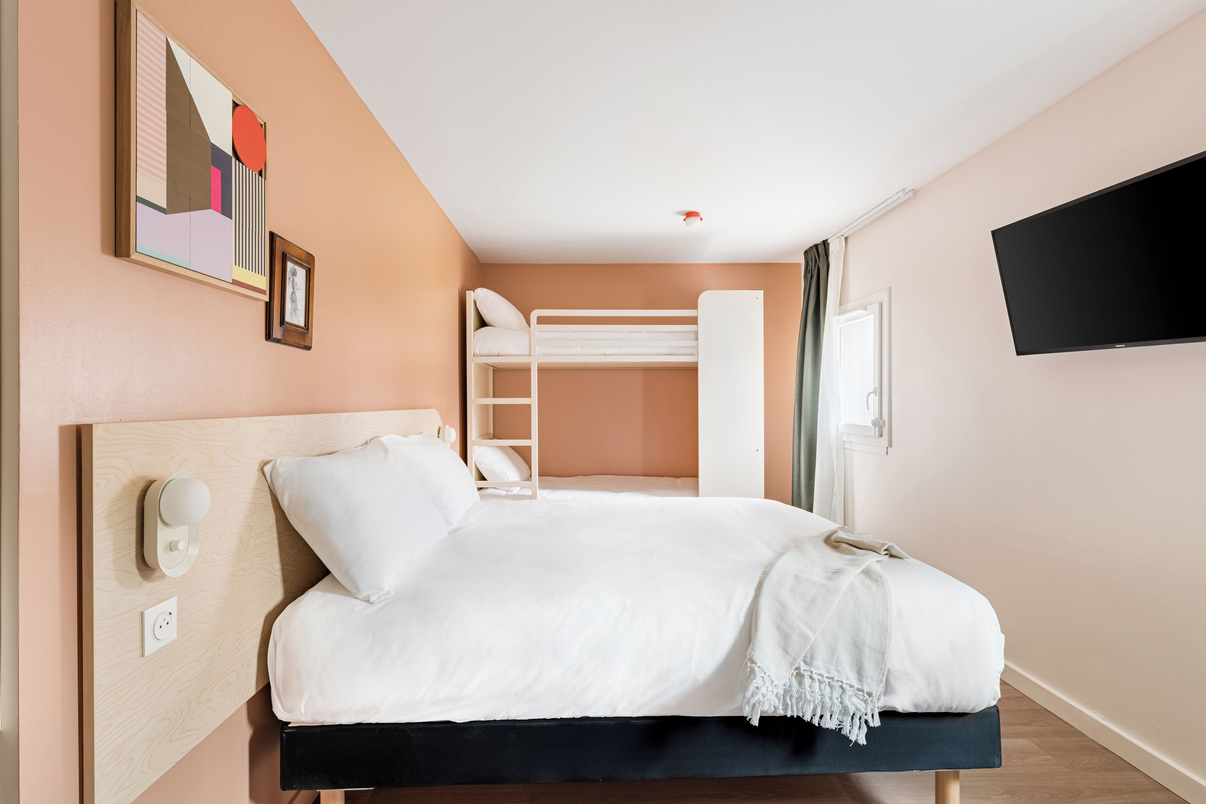 A hotel room with a bunk bed, a bedside table with a lamp, a TV mounted on the wall, and a painting on the wall.