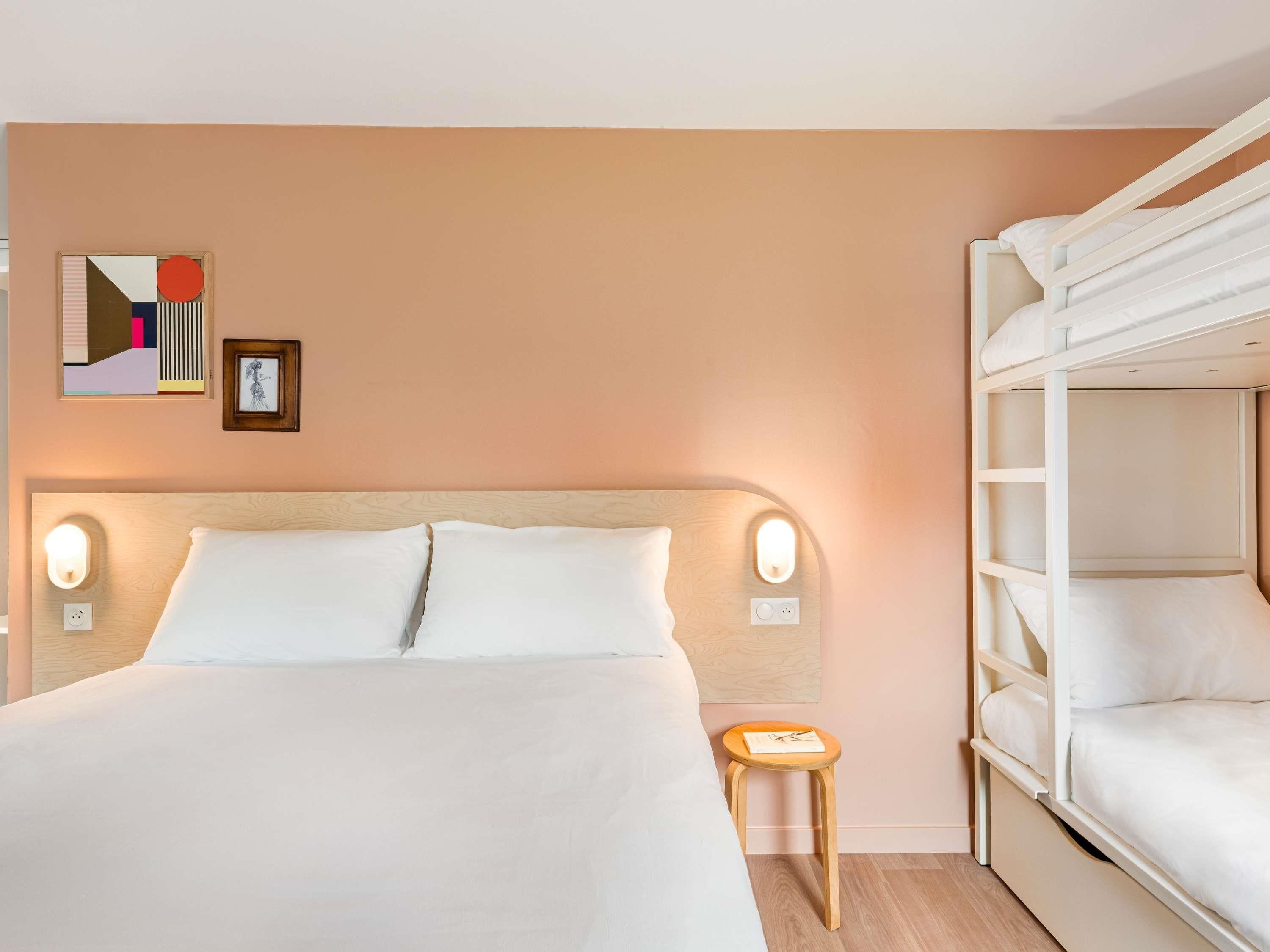 Une chambre d’hôtel avec un lit superposé, un grand lit avec deux oreillers, une table de chevet avec une lampe et un mur orné de tableaux.