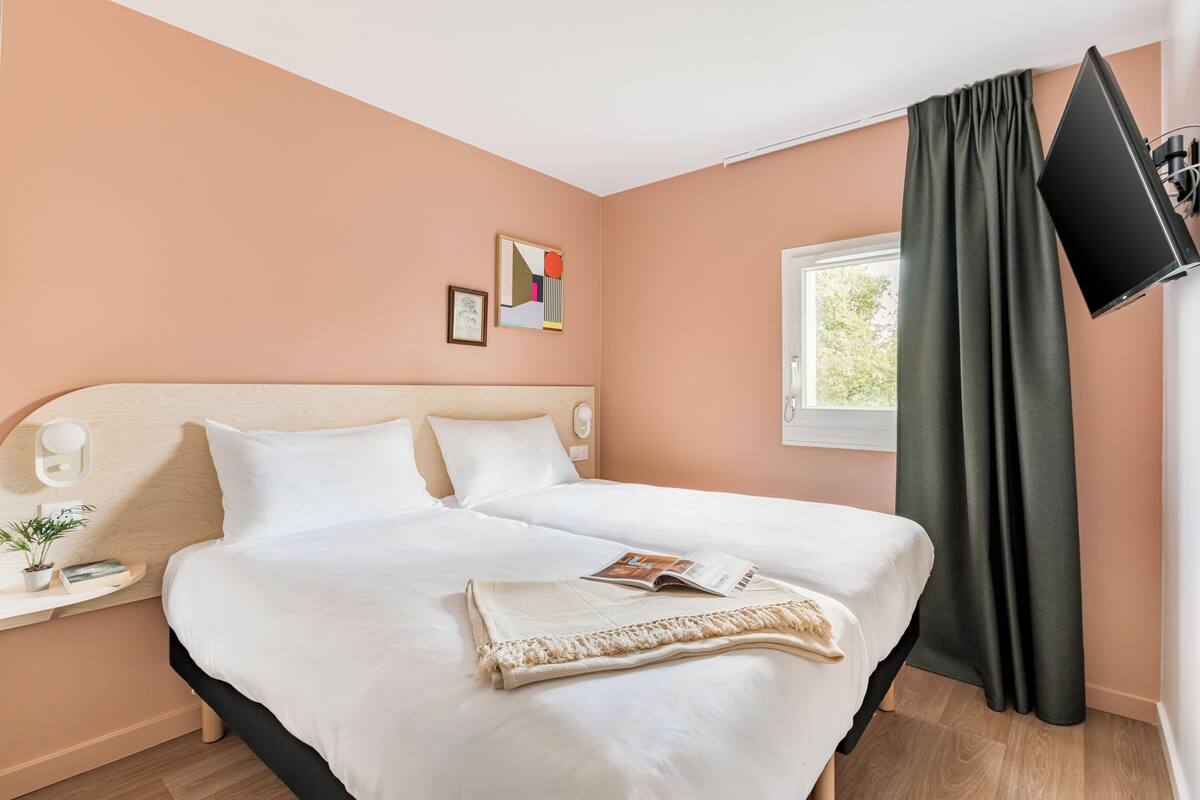A neatly made bed with white linens, a bedside table with a lamp, and a window with a dark curtain.