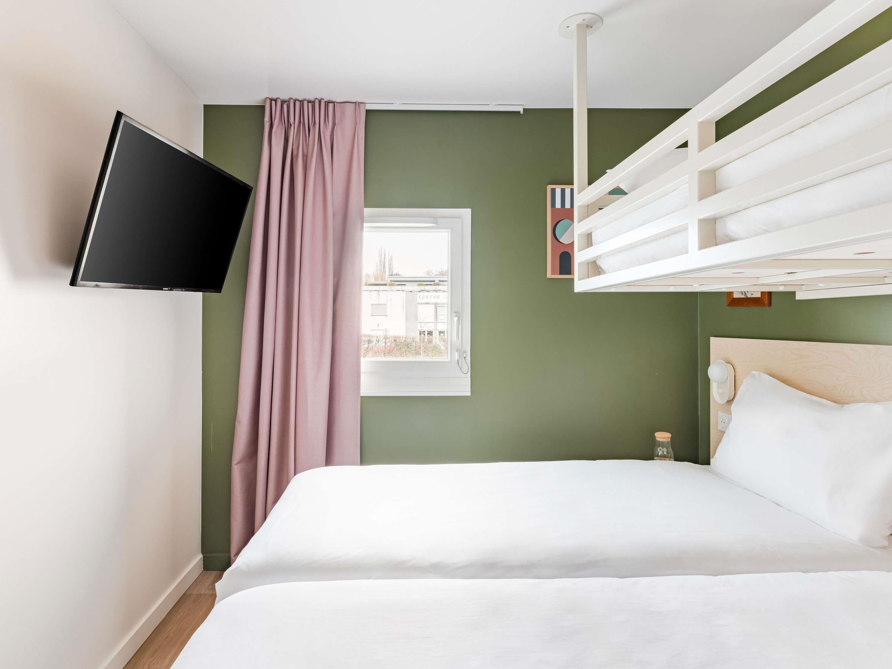 Une chambre avec un lit, un téléviseur fixé au mur, une fenêtre donnant sur des immeubles et une structure de lit superposé.