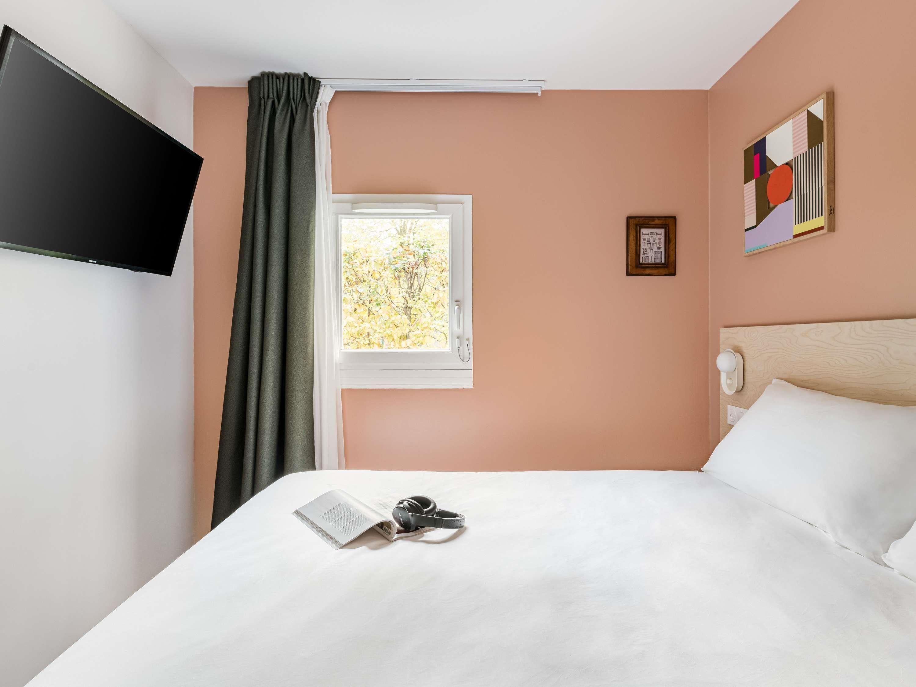 A hotel room with a bed, a television mounted on the wall, a window with curtains, and a painting on the wall.
