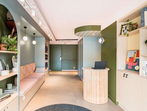 Un interior moderno con un mostrador de recepción curvo, una zona de estar con un banco y estanterías con plantas y libros.