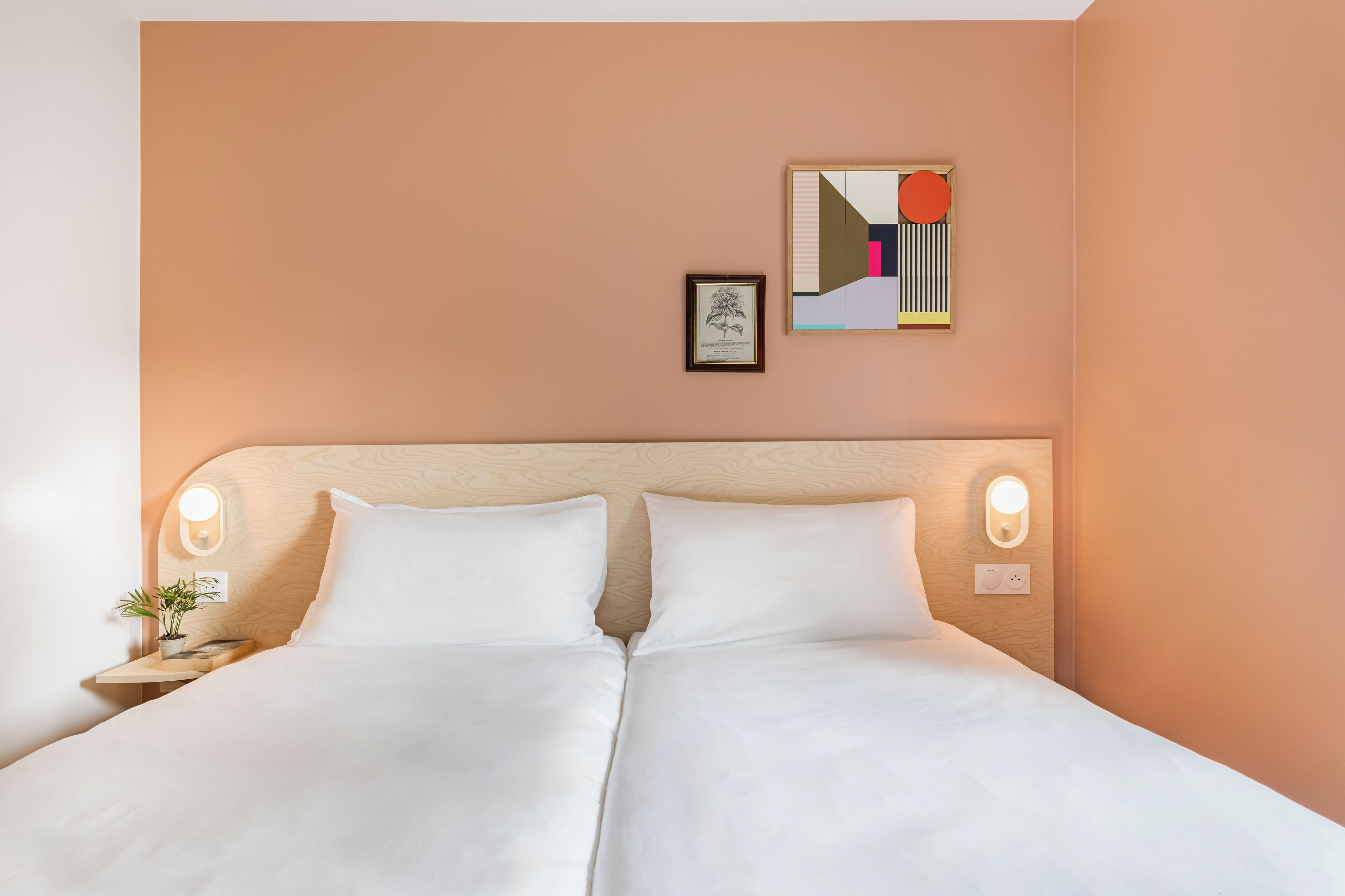 A double bed with white linens, a wooden headboard, bedside lamps, a small table with a plant, and a framed picture on the wall.