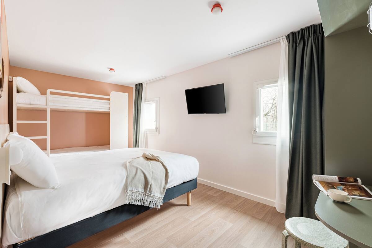 A room with a bunk bed, a flat-screen TV, a small table with a cup and a book, and a window with curtains.