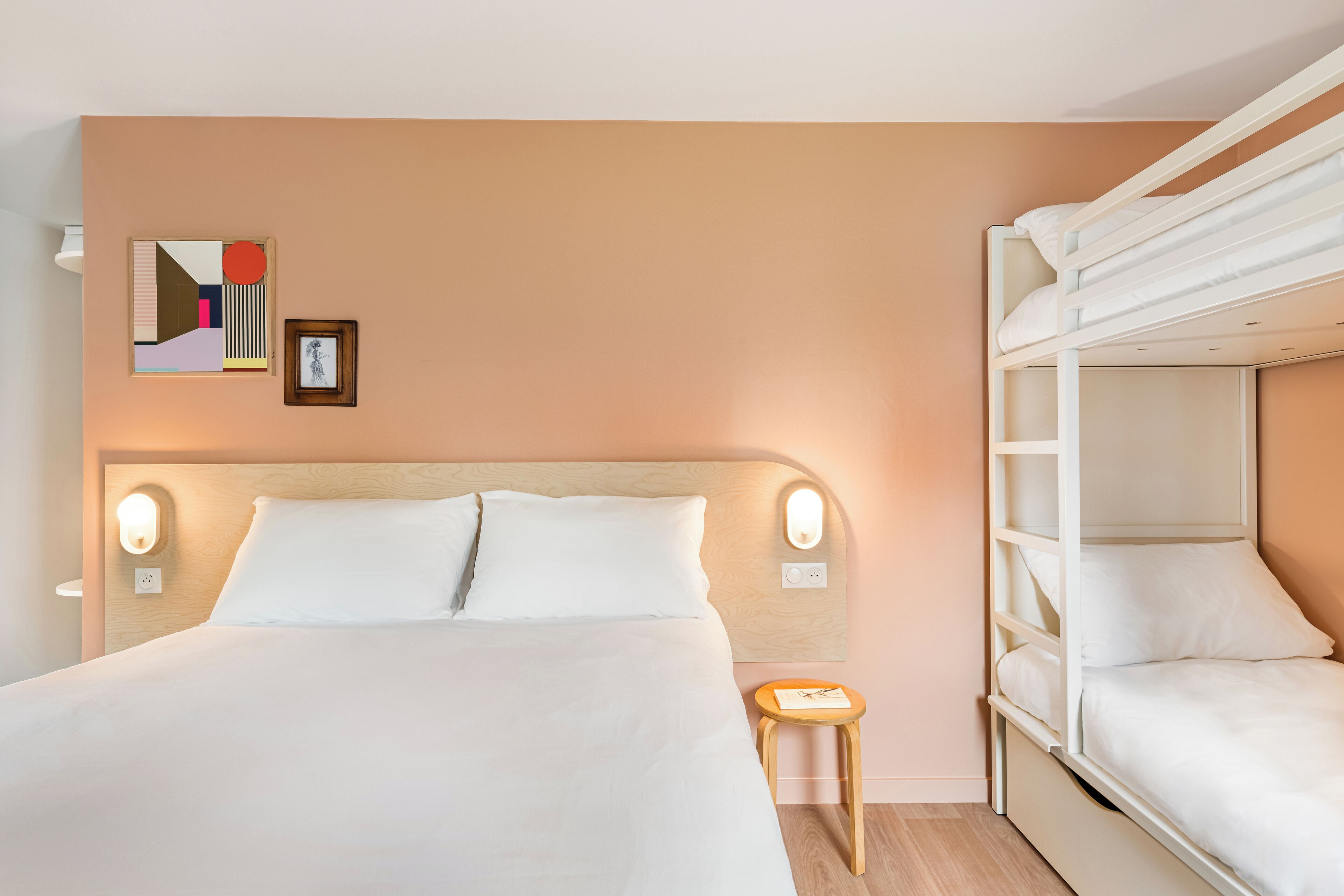 Habitación de hotel con una litera, una cama grande con ropa de cama blanca, una mesita de noche pequeña con una lámpara y una obra de arte colgada en la pared.