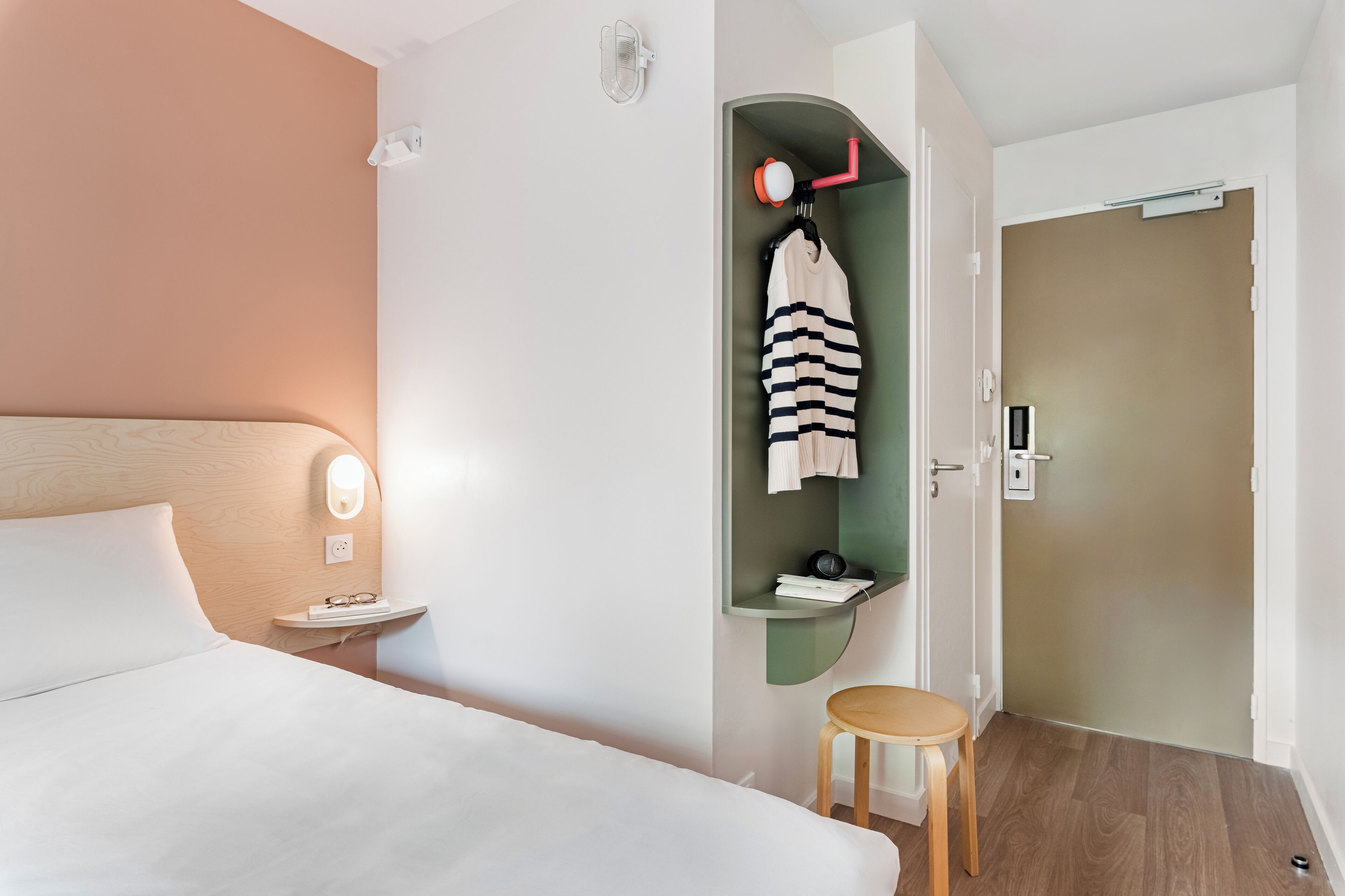 Una habitación de hotel con cama, mesita de noche, un taburete pequeño, un perchero con una prenda a rayas, un estante verde con un libro y un teléfono, y una puerta con cerradura.