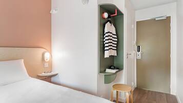 Una habitación de hotel con cama, mesita de noche, un taburete pequeño, un perchero con una prenda a rayas, un estante verde con un libro y un teléfono, y una puerta con cerradura.