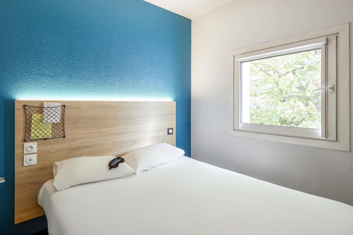 Ett hotellrum med en sänggavel i trä, en vit säng, ett fönster med utsikt över grönområden och ett eluttag.