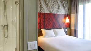 Camera d'albergo con doccia, un letto con lenzuola bianche, una testiera a motivi decorativi e una lampada da comodino.
