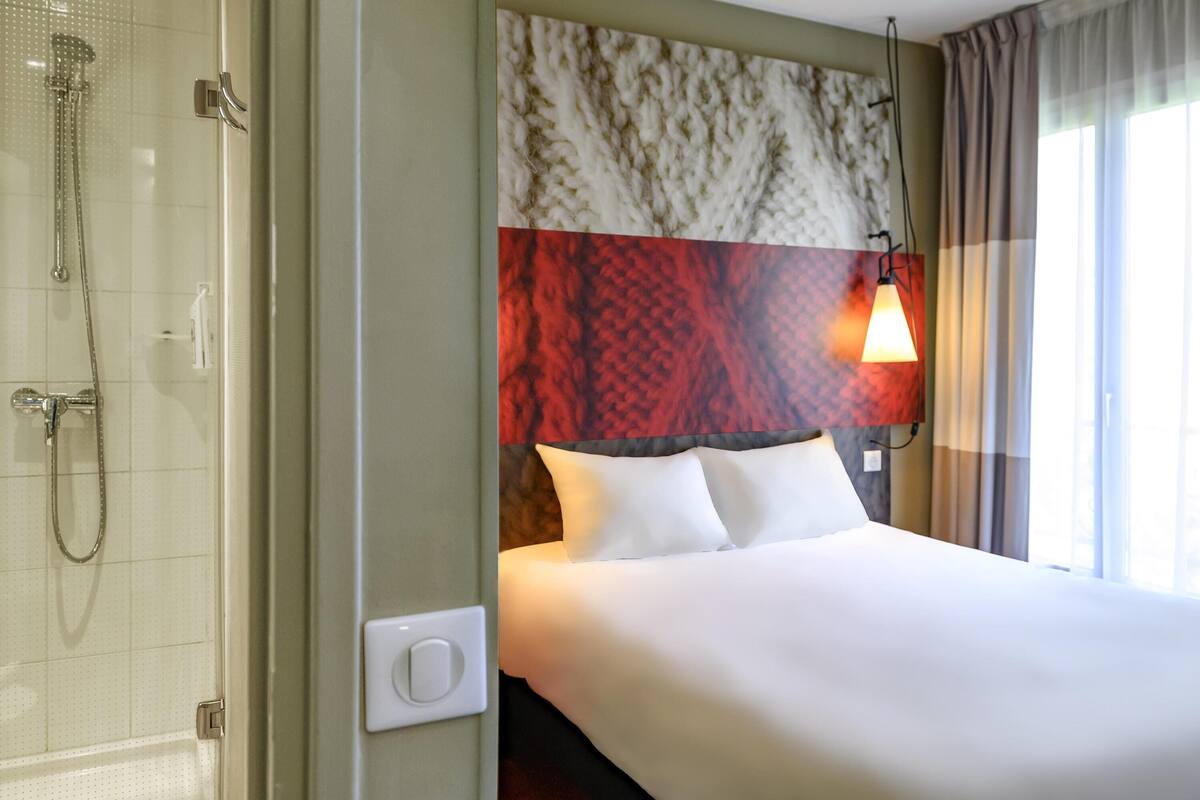 Habitación de hotel con ducha, cama con ropa blanca y lámpara de noche, además de un cabecero estampado.