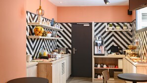A modern kitchen with a black and white chevron backsplash, wooden cabinets, and a black-framed door. There are two round tables with chairs, a hanging light fixture, and a shelf with fruits and condiments. - Ibis Paris Tour Montparnasse 15Ème (Paris)
