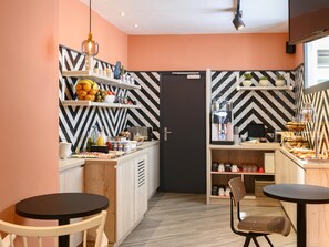 A modern kitchen with a black and white chevron backsplash, wooden cabinets, and a black-framed door. There are two round tables with chairs, a hanging light fixture, and a shelf with fruits and condiments. - Ibis Paris Tour Montparnasse 15Ème (Paris)