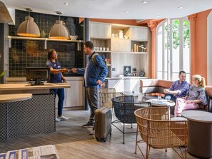 A modern hotel lobby with a reception desk, seating area, and a kitchen in the background.