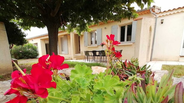 A garden with red flowers and green foliage in front of a building with a tiled roof and windows. - Josephine's House with swimming pool (Pierrelatte)