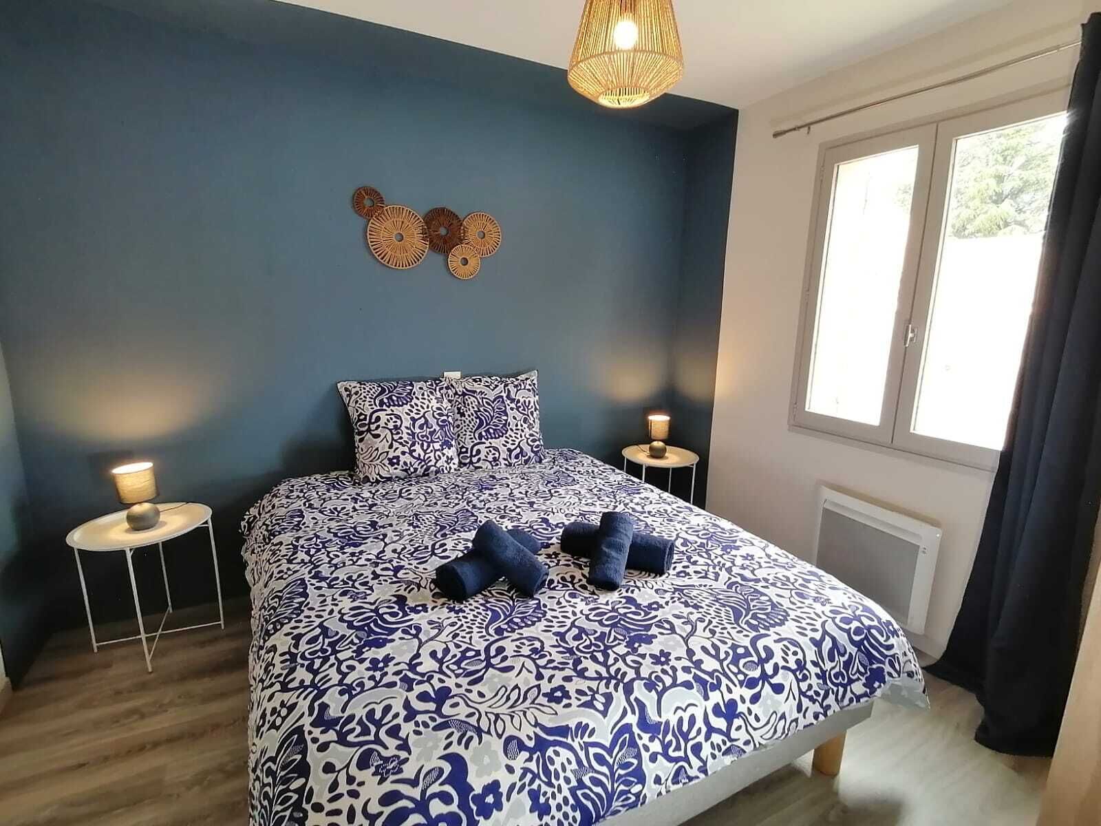 A bedroom with a blue and white patterned bedspread, two bedside tables with lamps, a decorative wall hanging, and a window with curtains.