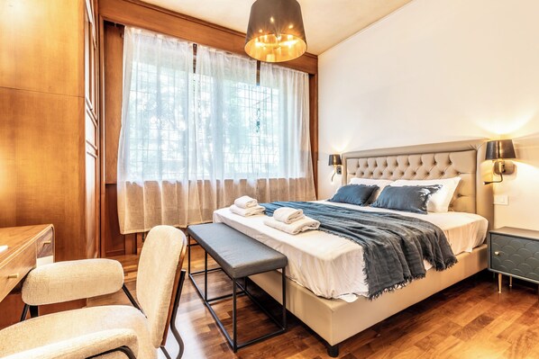 A bedroom with a bed, bench, chair, bedside table, lamp, and curtains. - Vatican SPA and Sauna Flatinrome (Rome)