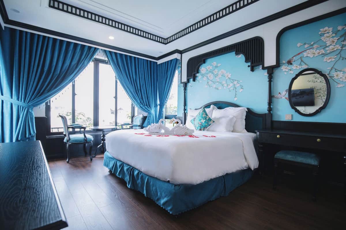 A bedroom with a large bed, a desk, and a chair. There is a window with blue curtains, a mirror on the wall, and a floral design on the ceiling.