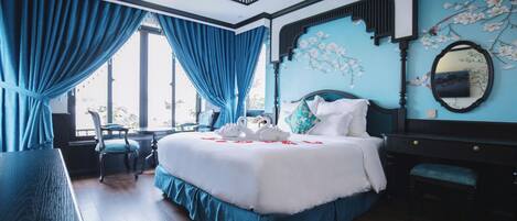 A bedroom with a large bed, a desk, and a chair. There is a window with blue curtains, a mirror on the wall, and a floral design on the ceiling.