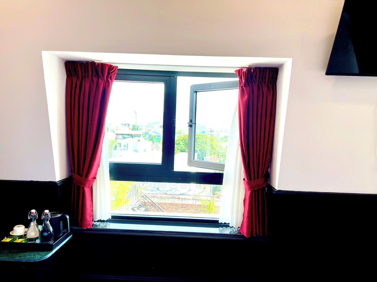 A room with a window featuring red curtains, a black frame, and a view of buildings and greenery outside.