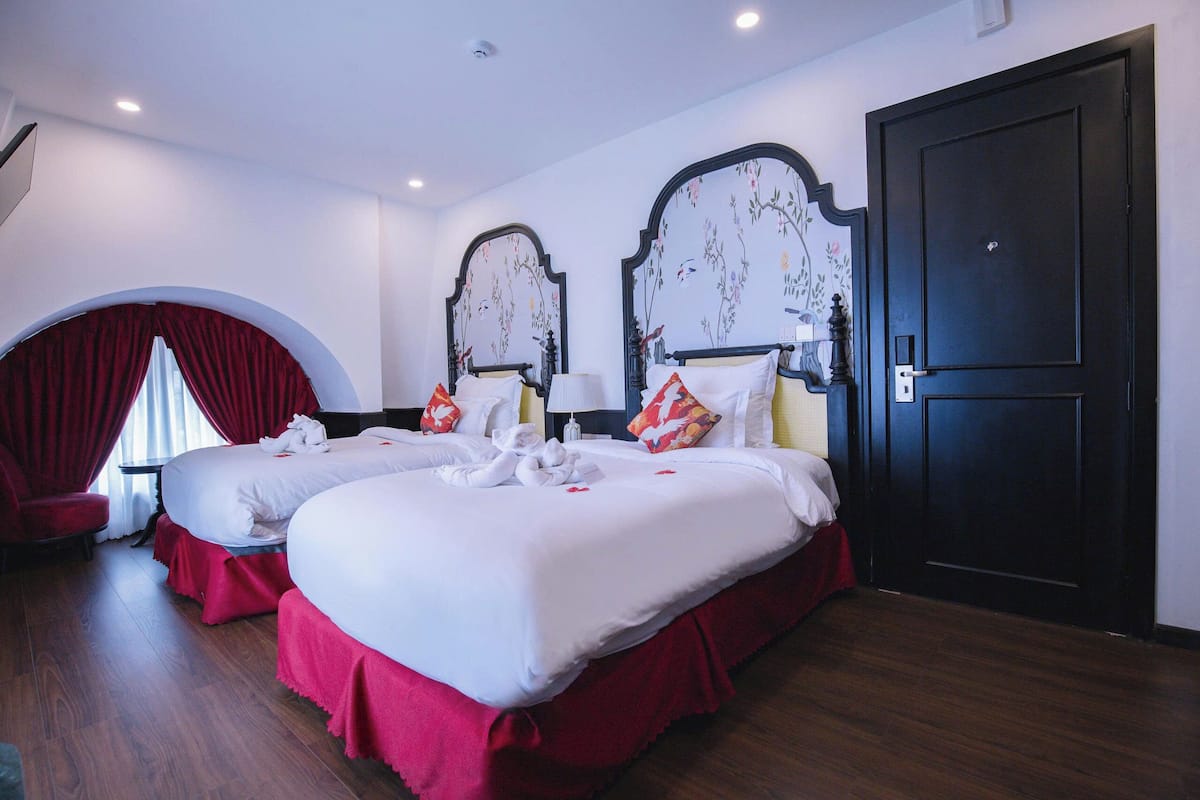 A hotel room with two beds, red curtains, a black door, and a small table with a lamp.