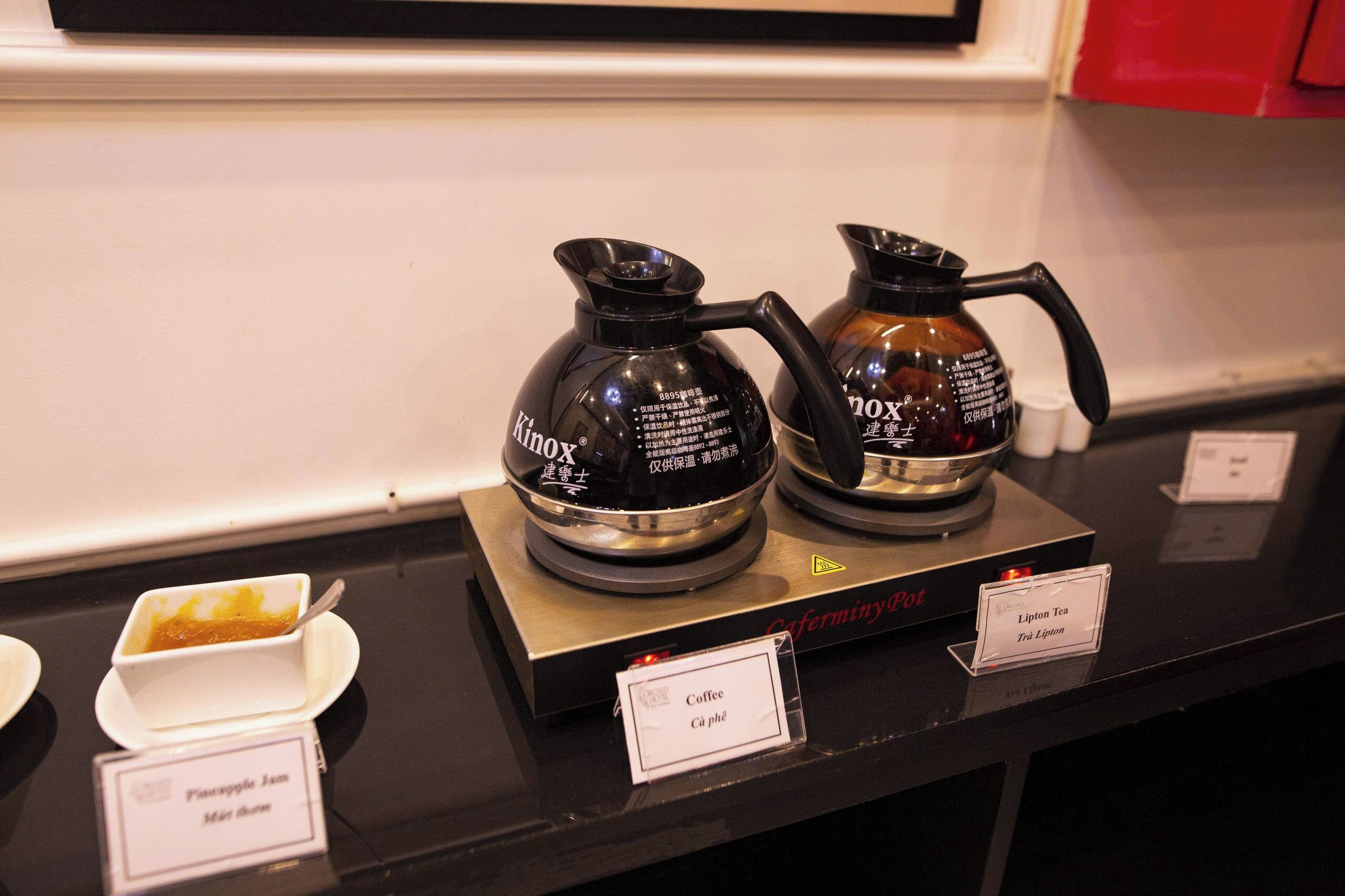 Una estación de café con dos máquinas de café identificadas como 'Kino' y 'Nox' sobre un mostrador. Hay tazas pequeñas con café y un letrero que indica 'Café' y 'Cà phê'.