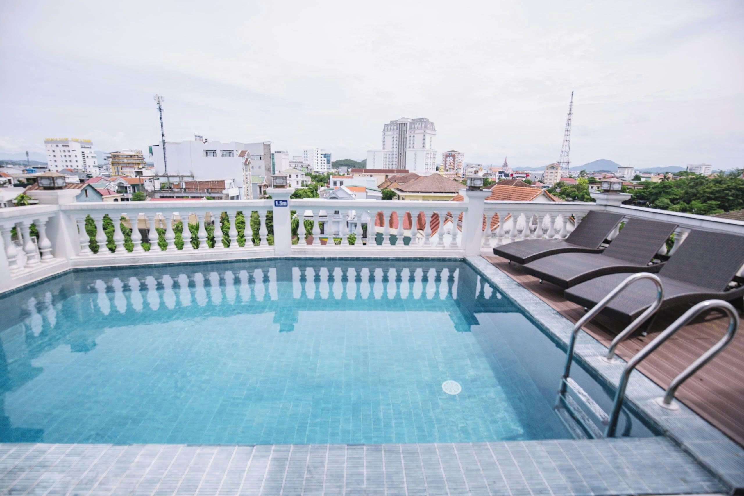 A rooftop pool with a white balustrade, lounge chairs, and a cityscape view in the background.