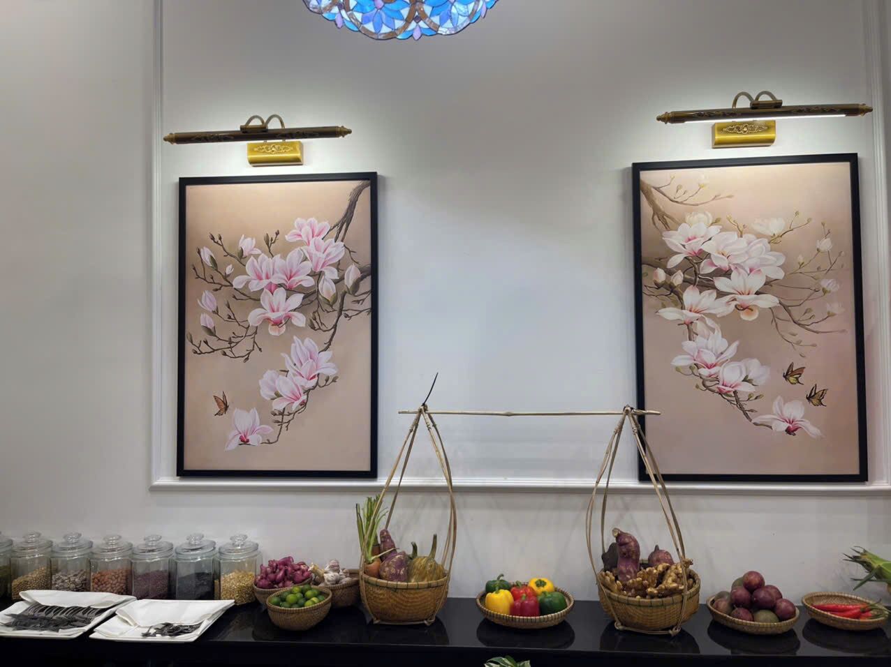 Deux tableaux représentant des motifs floraux, encadrés et accrochés au mur, une fenêtre donnant sur un paysage, et une table sur laquelle sont disposés des paniers remplis de fruits et de légumes.
