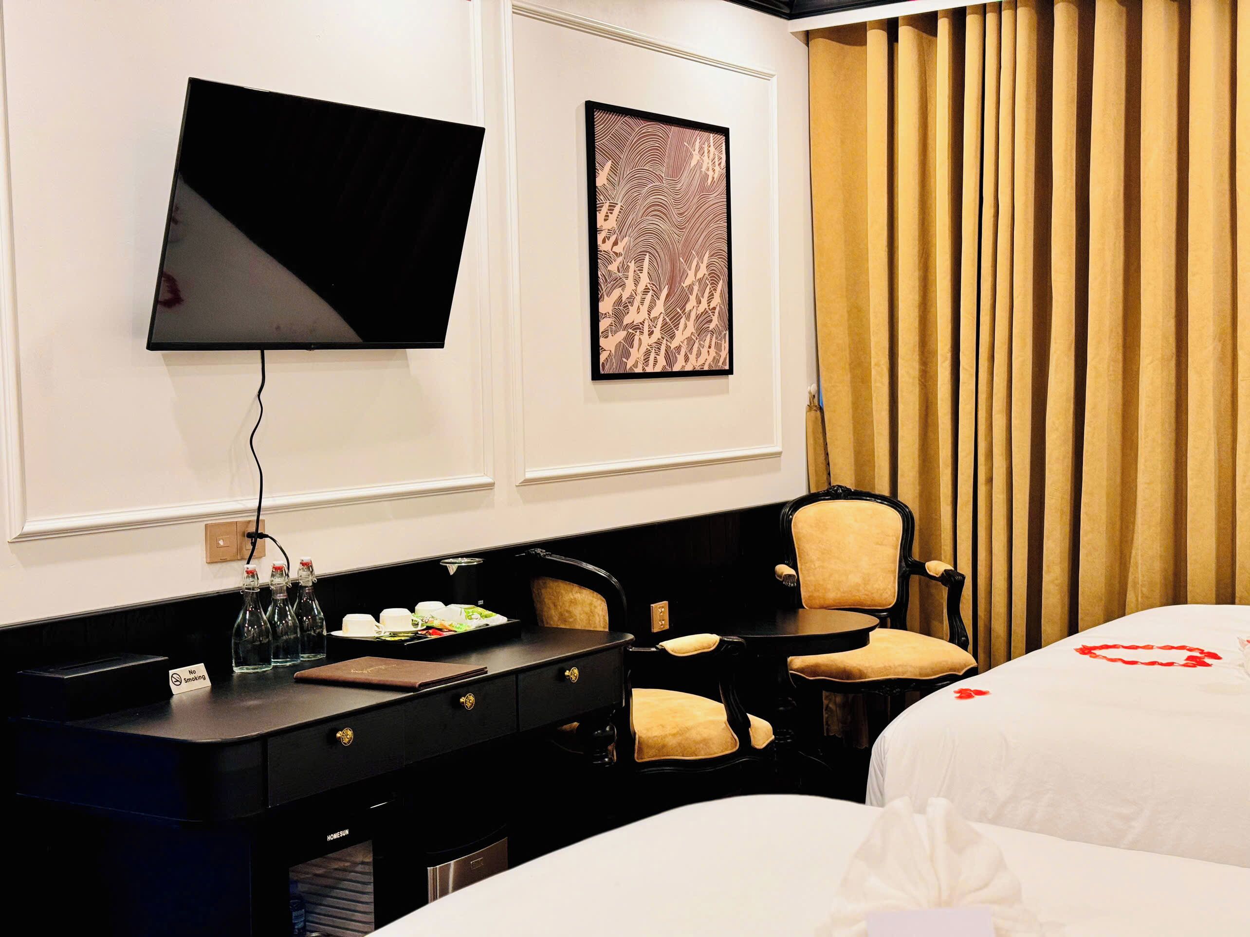 A hotel room with a flat-screen TV, a desk with a coffee machine, a chair, and a bed with a red heart decoration.