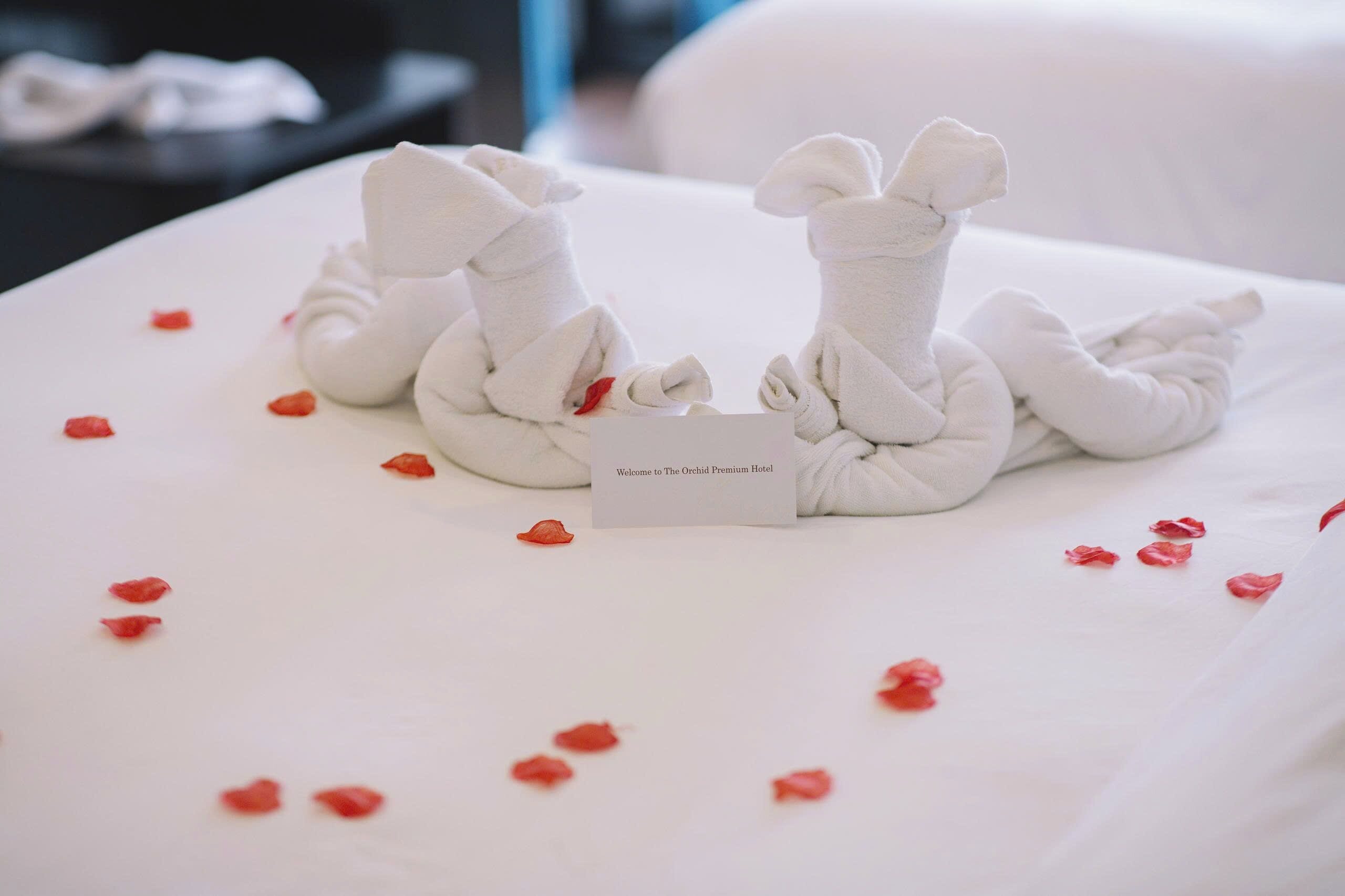 A bed with a towel arrangement forming a bow, a welcome card, and scattered rose petals.