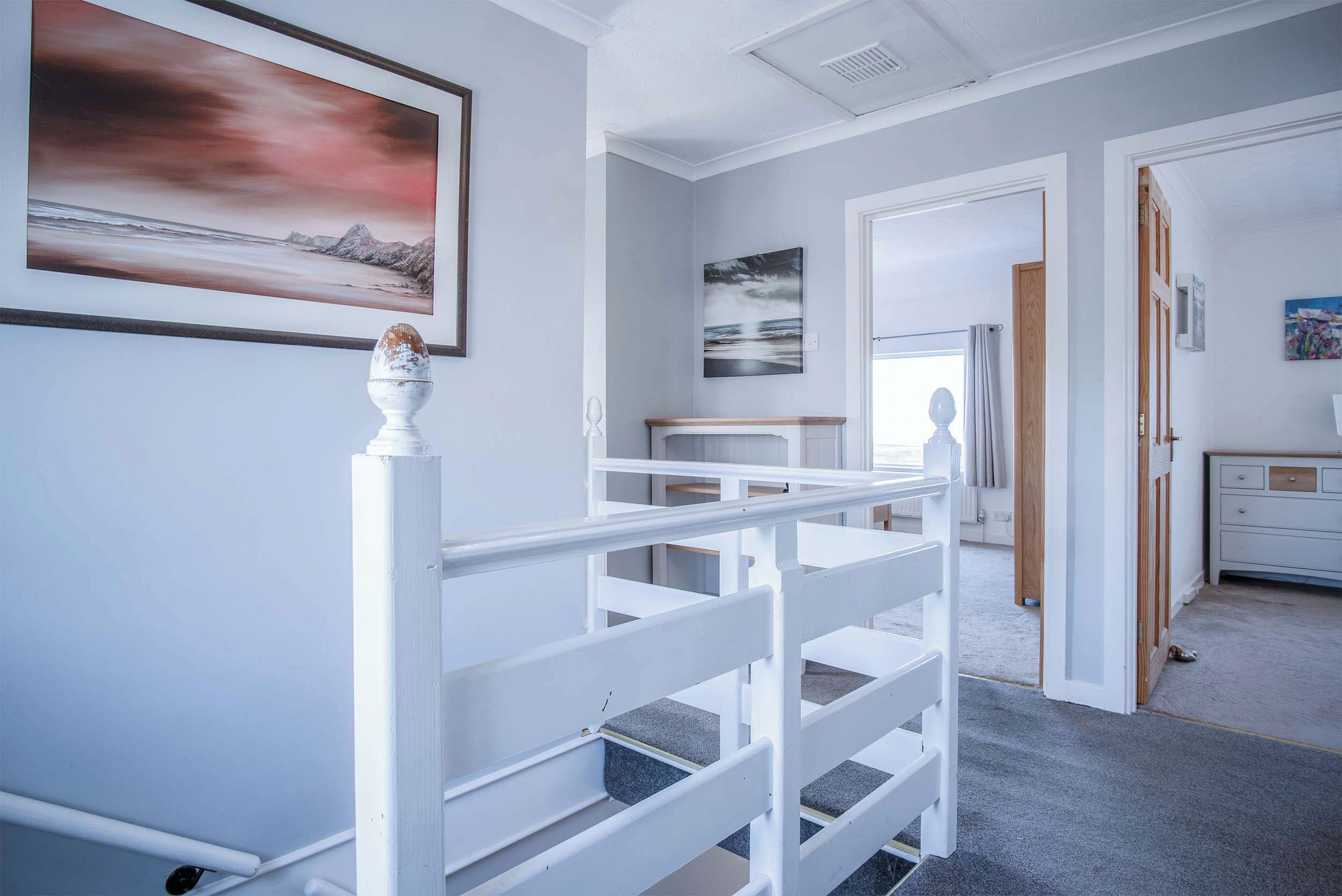 Un escalier avec des rampes blanches mène à une pièce où se trouvent un lit, une commode et un tableau représentant une scène de plage.