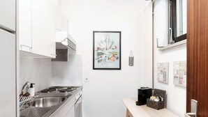 A compact kitchen with white cabinets, a stainless steel sink, and a stove. There's a framed artwork on the wall and a small table with a basket.