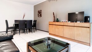 A modern living room with a glass coffee table, a sofa, a dining area with chairs, a wooden cabinet, and a flat-screen TV.