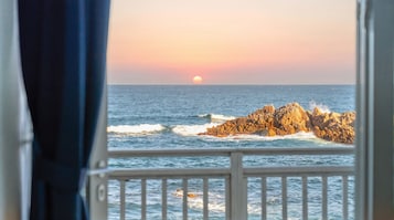 Vista del océano con olas rompiendo contra afloramientos rocosos, un atardecer al fondo y una ventana con marco blanco.