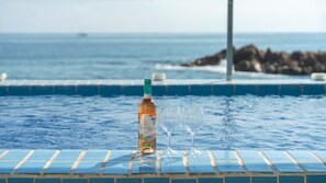 A bottle of Moulon Cadet 2022 wine and two glasses on a tiled pool edge with the ocean and rocky shore in the background. - Yeongdeok Chaeumneul Poolvilla (Yeongdeok)