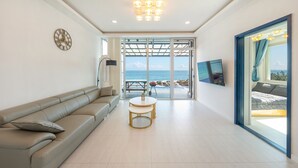 Een moderne woonkamer met een beige bank, een ronde tafel en een groot raam met uitzicht op zee.