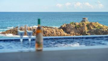 A rocky island with a small structure, a pool with two glasses and a bottle, and a view of the ocean.