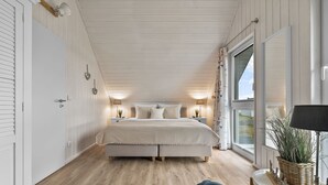 Et soveværelse med en stor seng, trægulv, et vindue med gardiner og et natbord med en lampe.