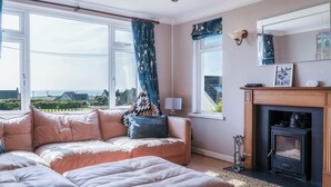 A living room with a sofa, ottoman, and a fireplace. - Carissima - 4 Bedroom House - Rhossili (Swansea)