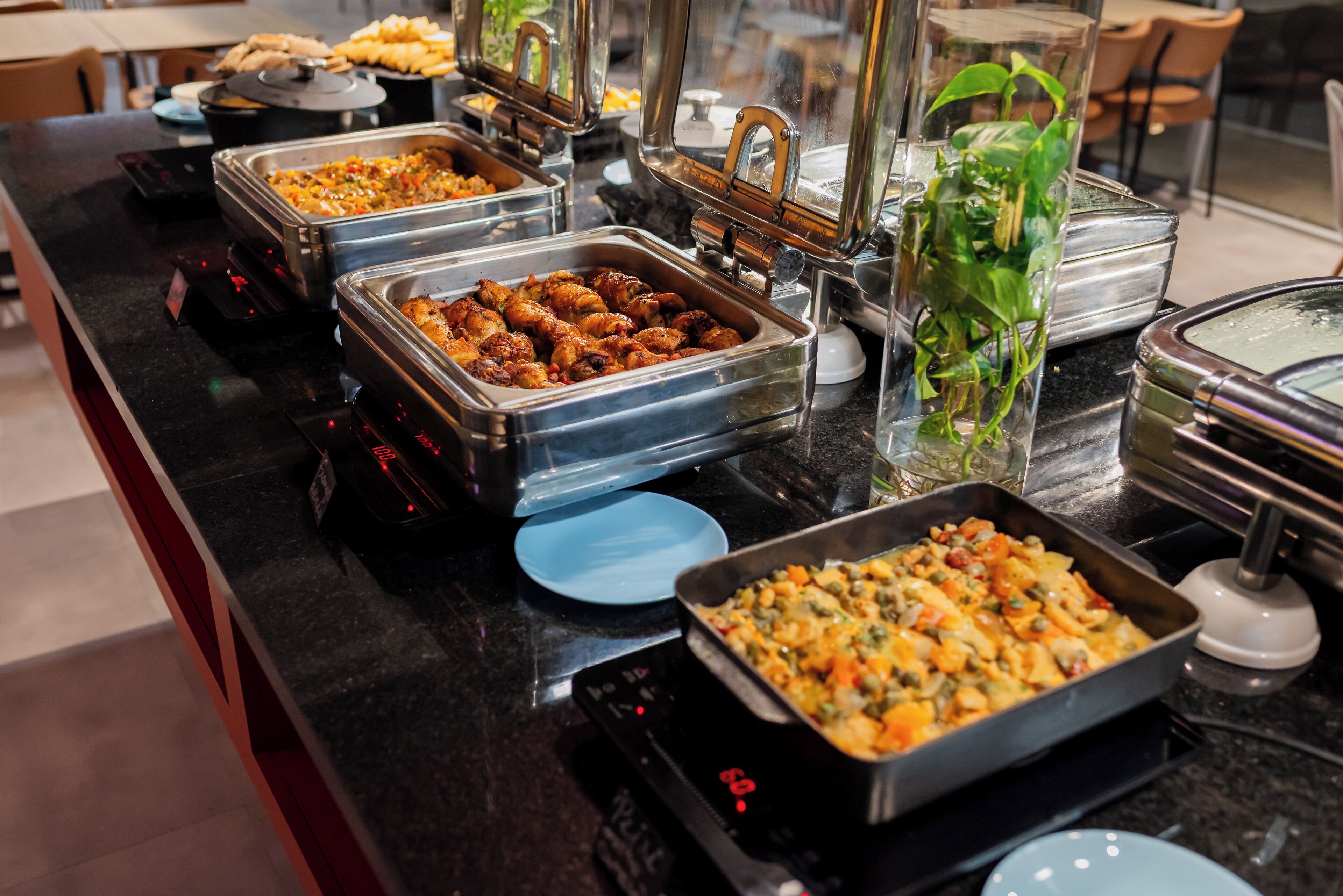 A buffet with various dishes in metal containers, a glass vase with green plants, and a digital scale.