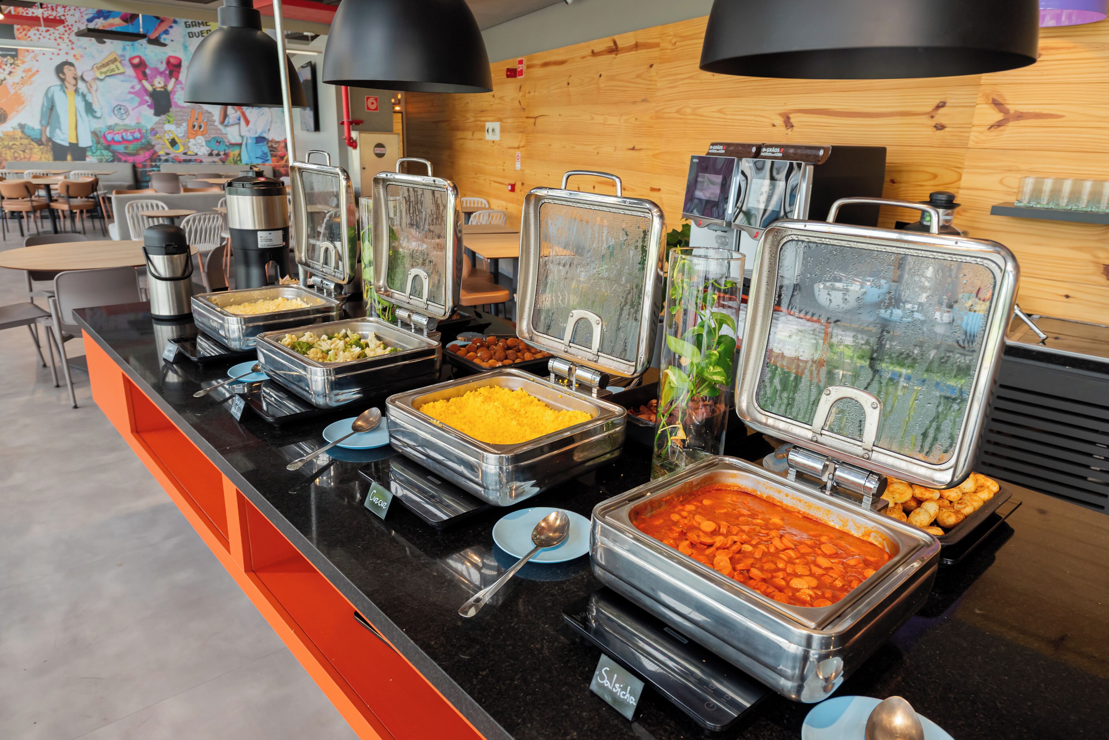 Un bufé con diferentes alimentos en recipientes metálicos, una estación de café y una zona de estar con sillas y mesas.