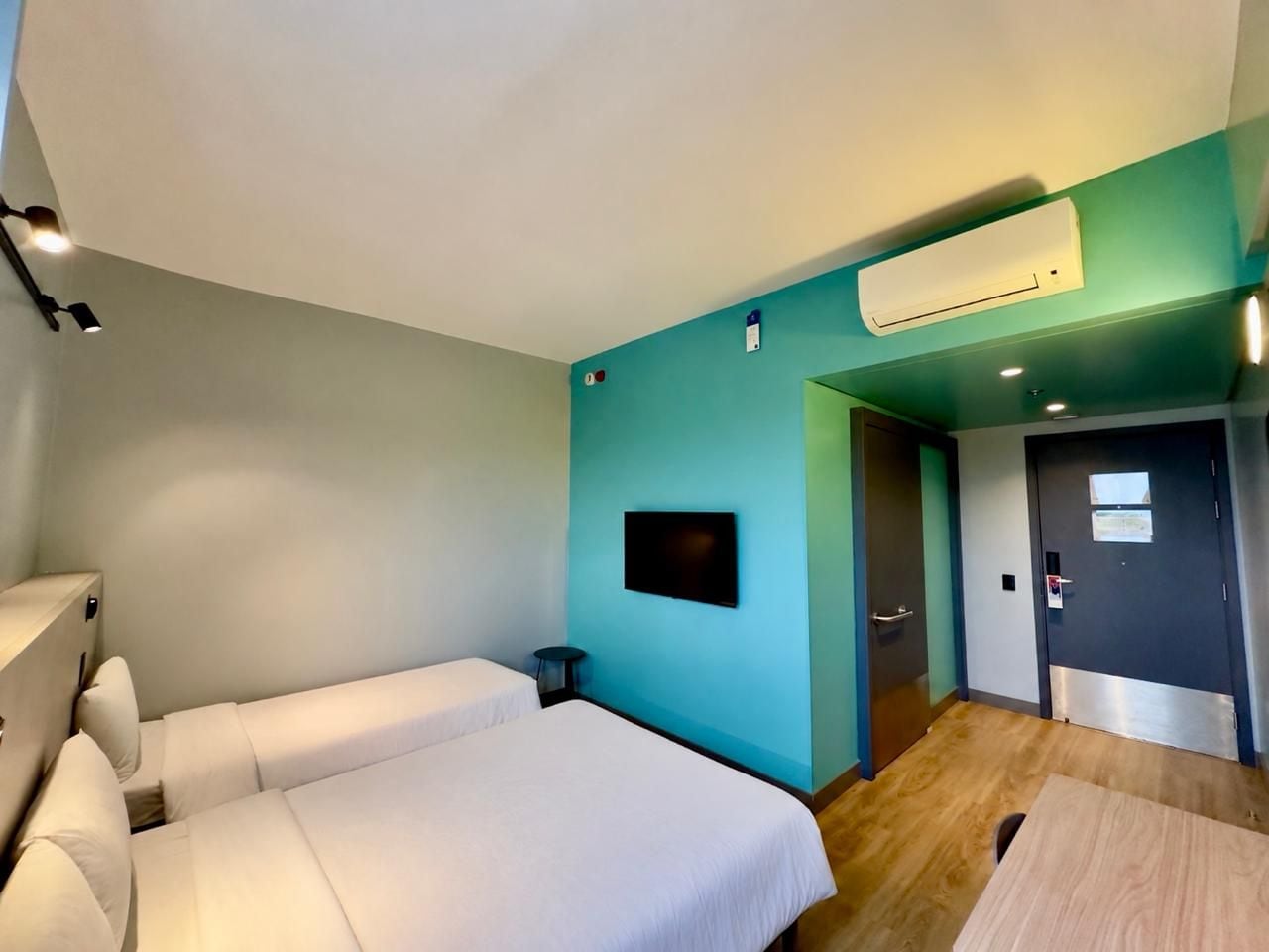Una camera d'albergo con due letti, una televisione, un condizionatore d'aria e una porta che dà su un corridoio.