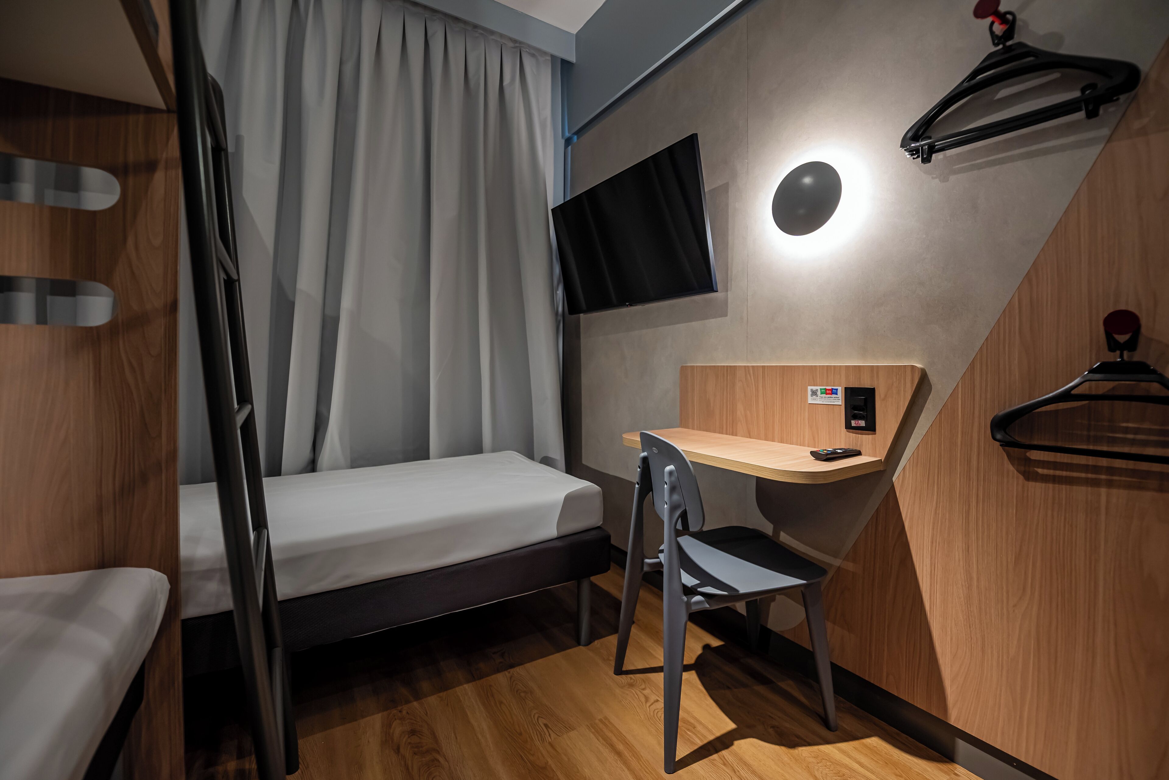 A compact hotel room with a bunk bed, a desk with a chair, a flat-screen TV, and a coat rack.
