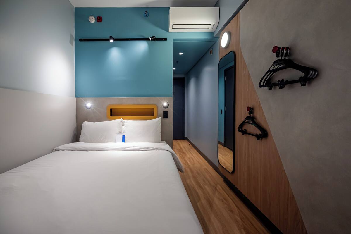 Una habitación de hotel con una cama, cabecera de madera, un espejo y un perchero con ganchos.