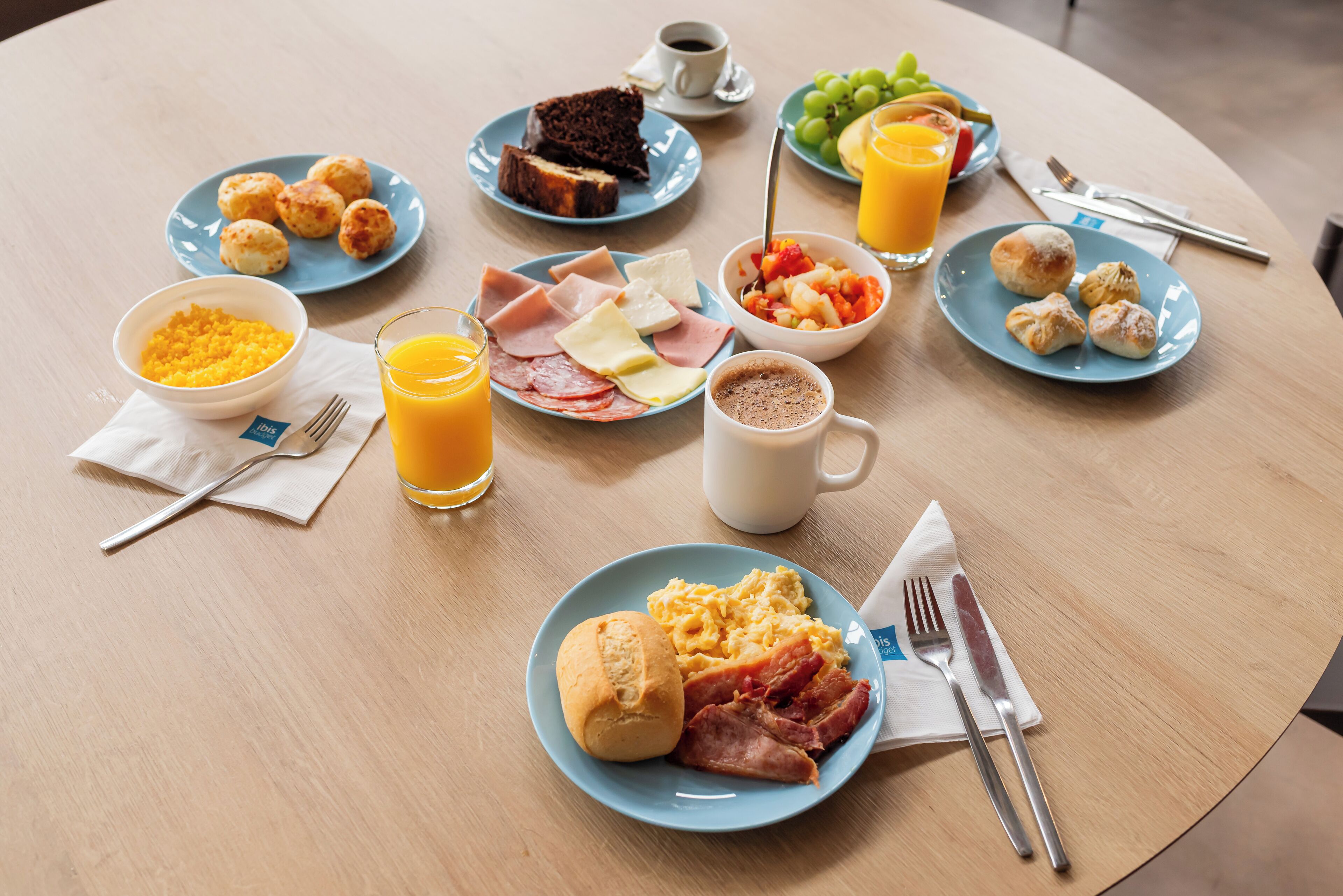 A breakfast table with plates of eggs, ham, cheese, and pastries, along with a bowl of fruit and a glass of orange juice.