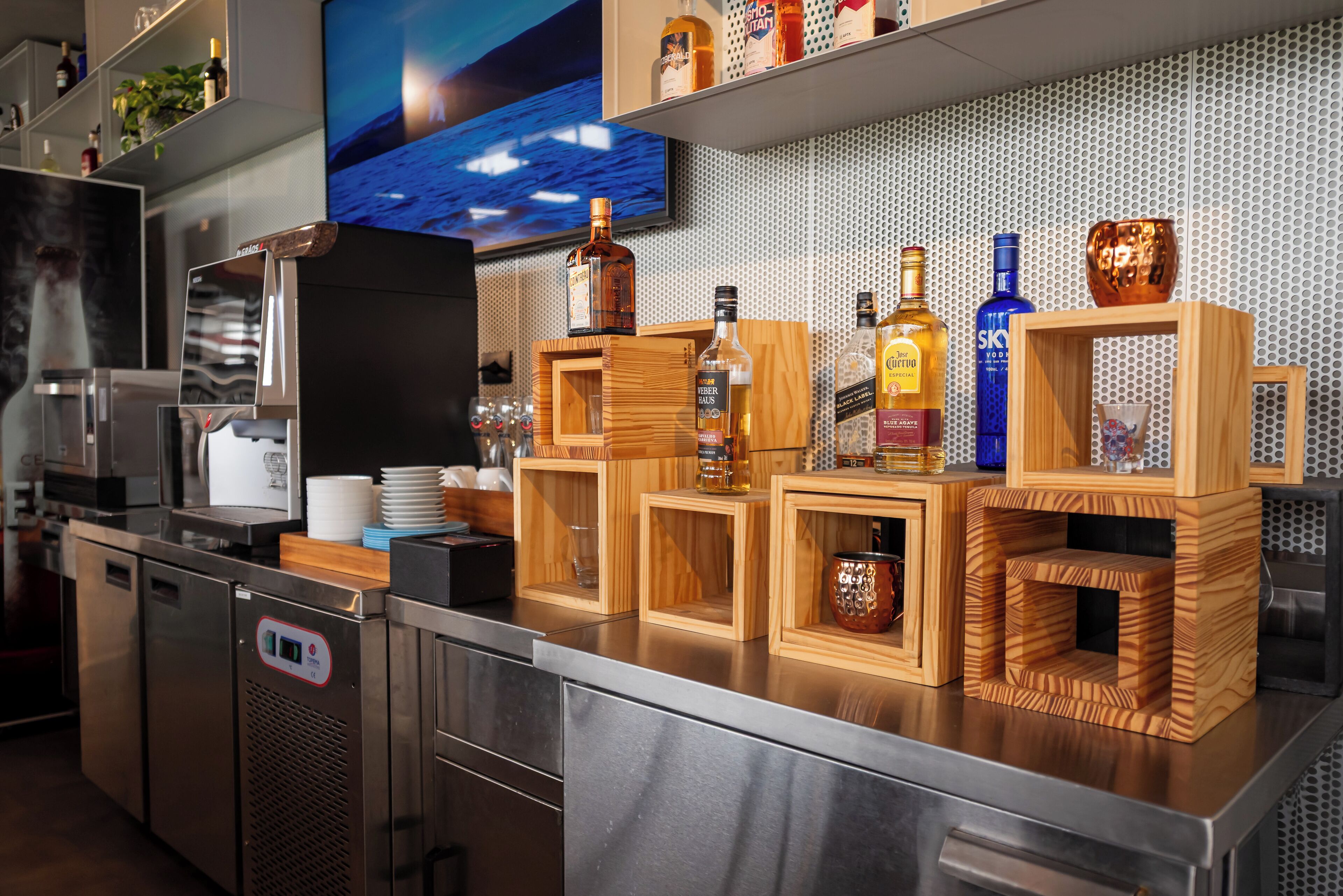 A bar area with wooden shelves holding various bottles, a television mounted on the wall, and a stainless steel refrigerator.