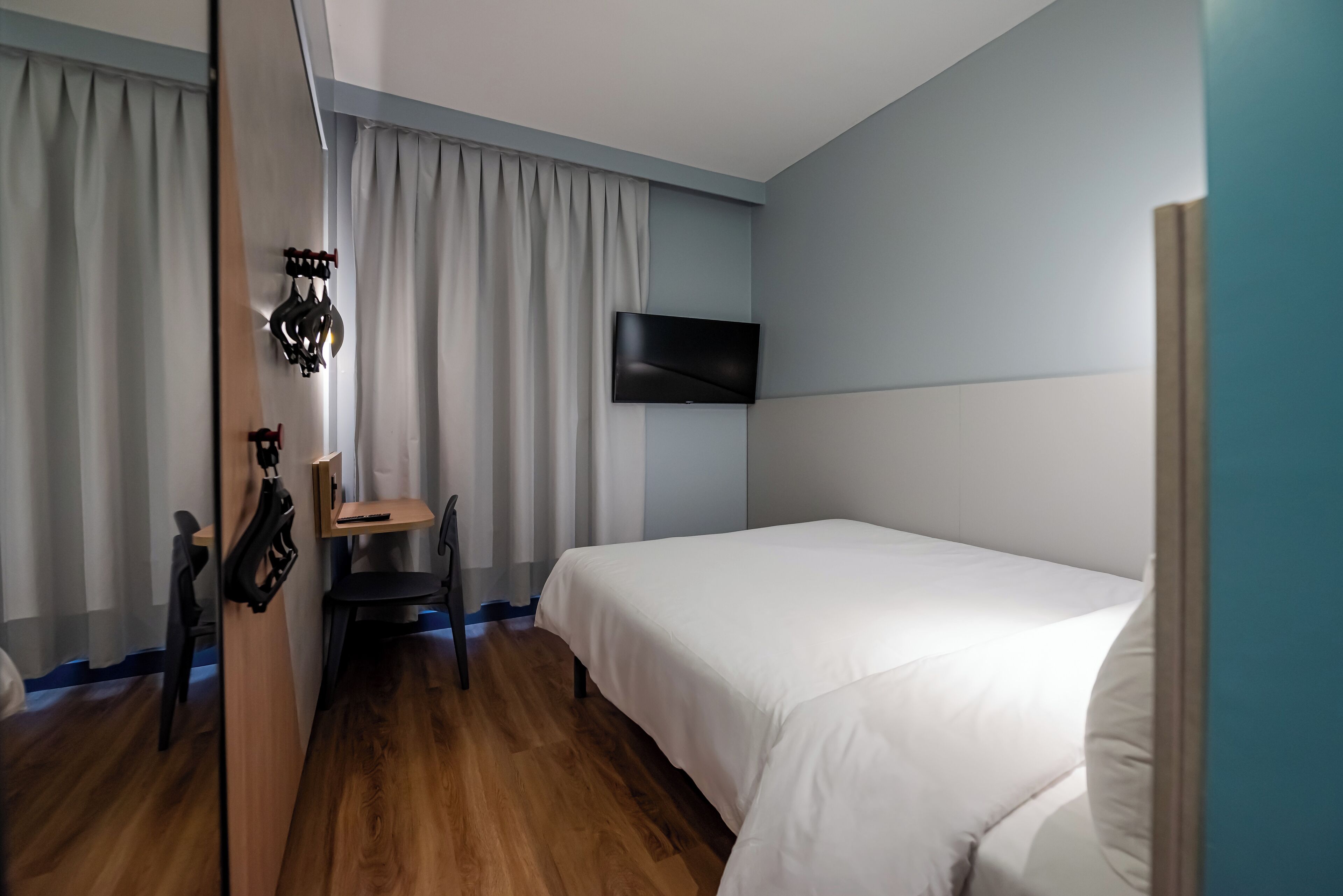Quarto de hotel com piso de madeira, cama com roupa de cama branca, escrivaninha com cadeira, TV de tela plana na parede e janela com cortinas.