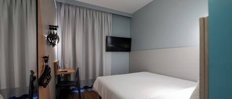 A hotel room with a wooden floor, a bed with white bedding, a desk with a chair, a flat-screen TV mounted on the wall, and a window with curtains.